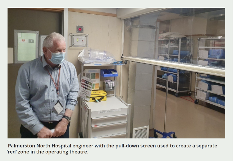 Palmerston North Hospital engineer with the pull-down screen used to create a separate ‘red’ zone in the operating theatre. The image shows the engineer wearing a blue medical mask, and the clear plastic screen is partially pulled down so that the viewer can tell that it is not a permanent barrier.